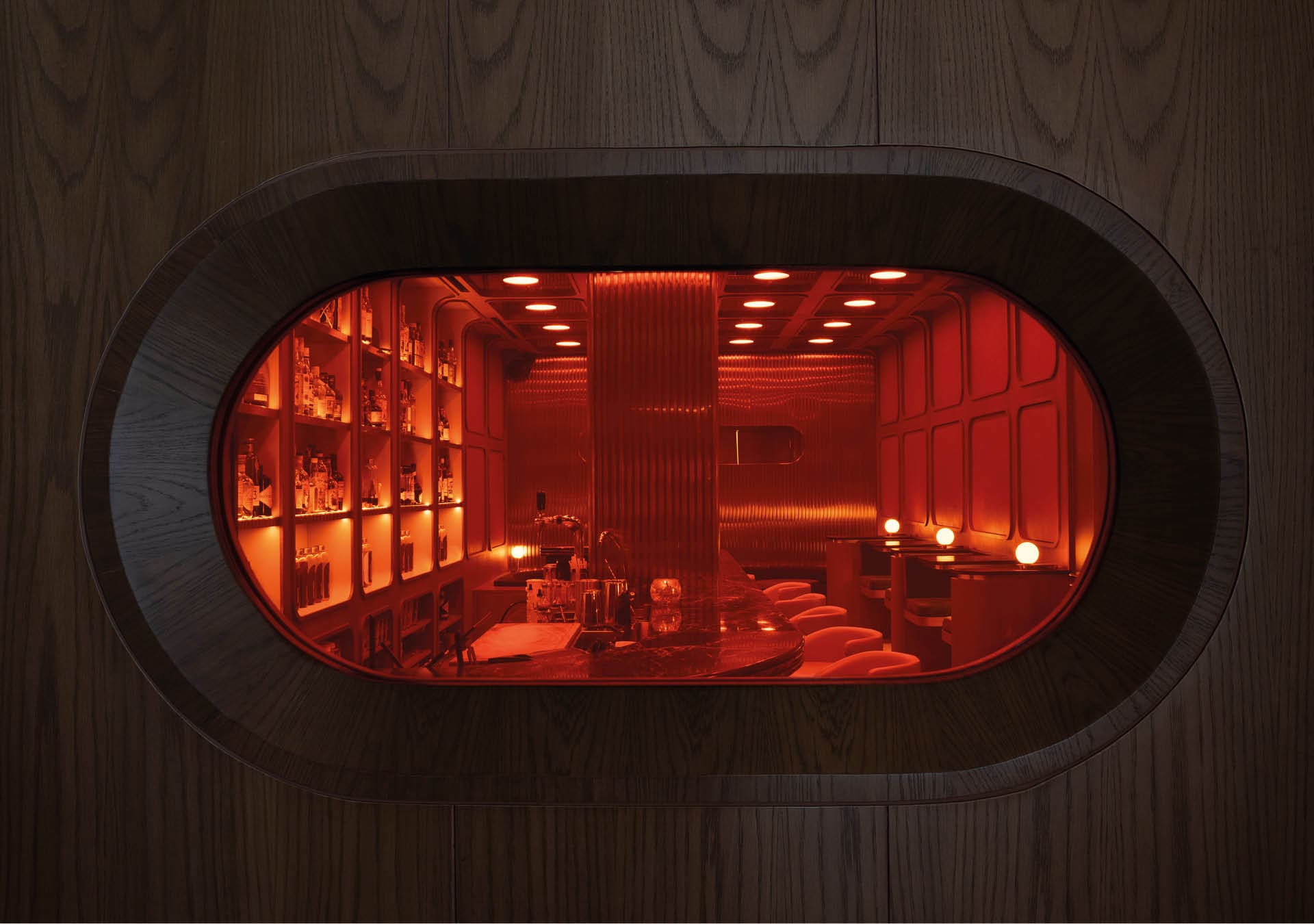 A futuristic oblong-shaped window looking into the red-lit interiors of Night Hawk cocktail bar in Singapore.