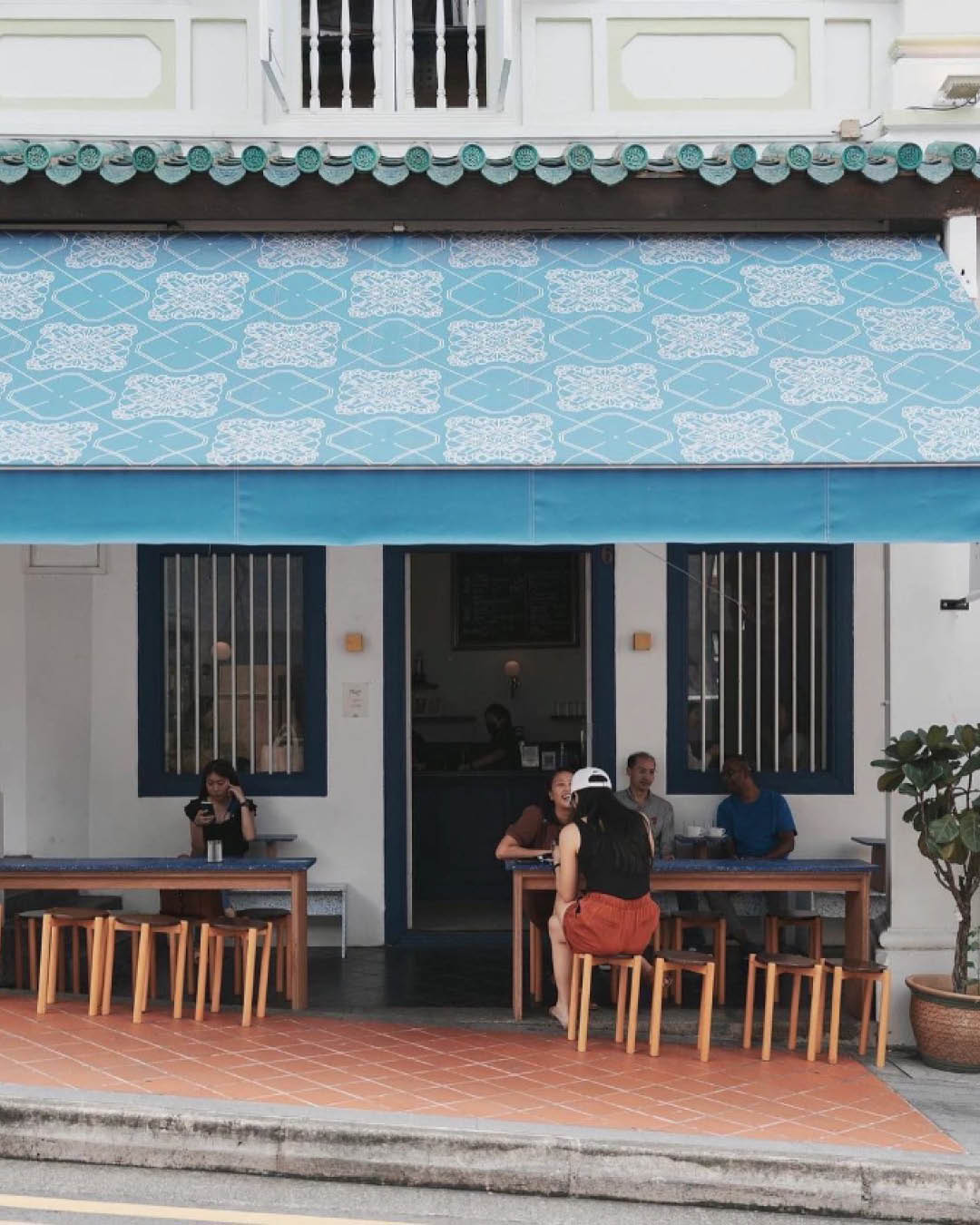 A street view of Maxi Coffee Bar, where two tables with stalls sit under a blue awning.