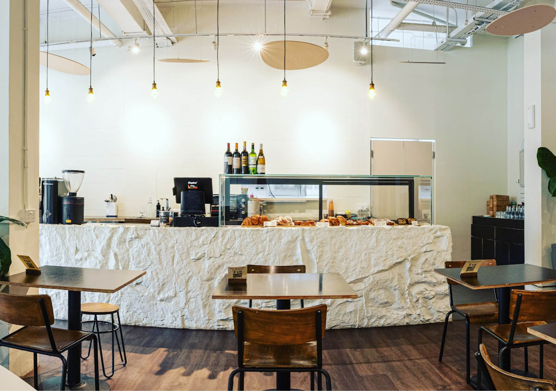 A white counter with pastries and coffee machines at the back of Twenty Eight Cafe in Singapore with wooden chairs and tables in front of it.
