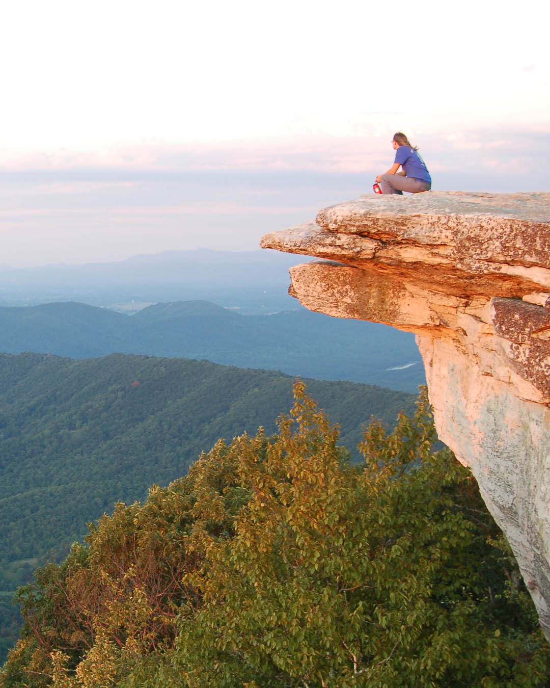 A person sits on the top of a rocky promontory gazing out over tree-covered rolling hills below.