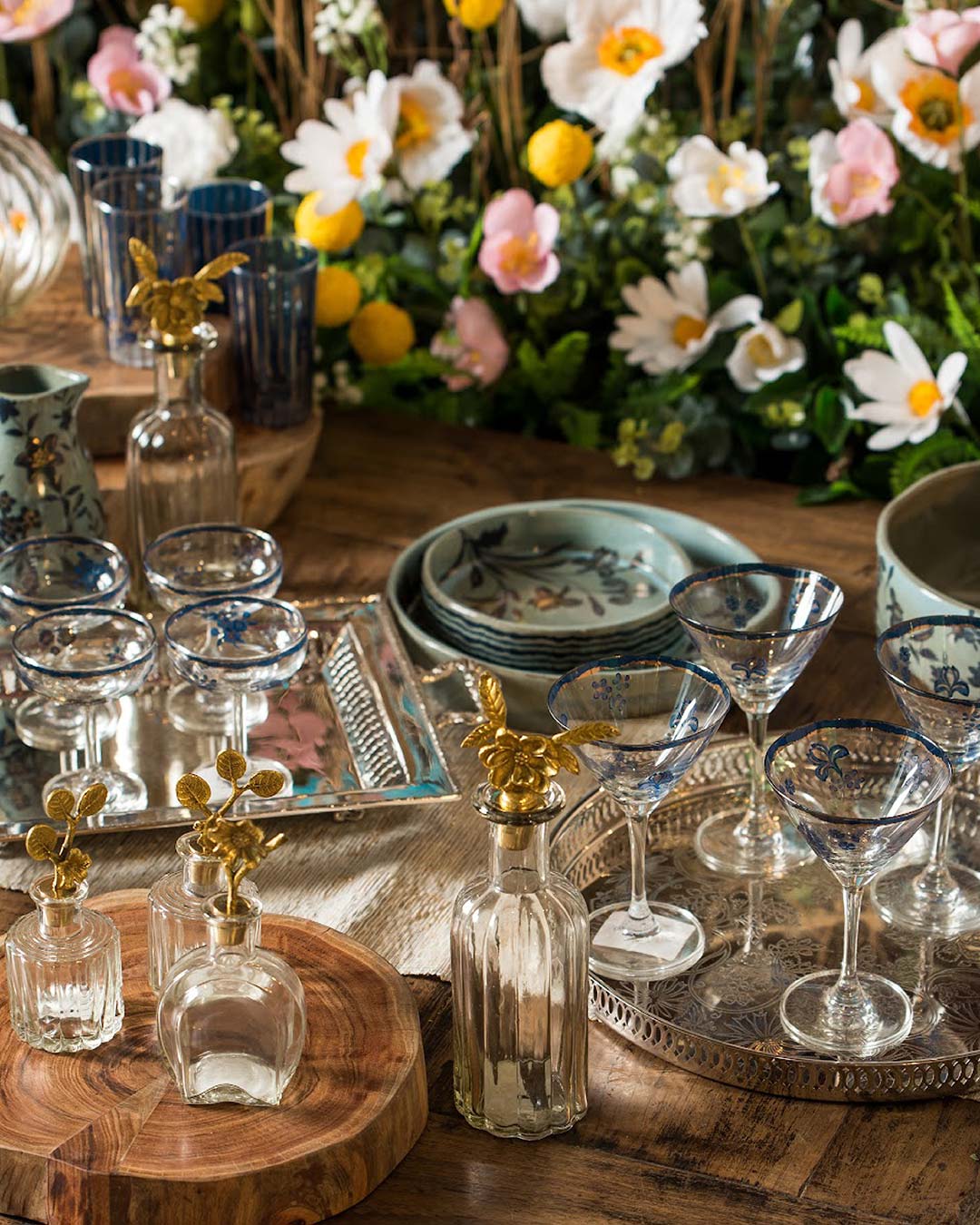 Glasses, bowls and other tableware arranged on a wooden table in front of flowers at Good Earth boutique in Mumbai.