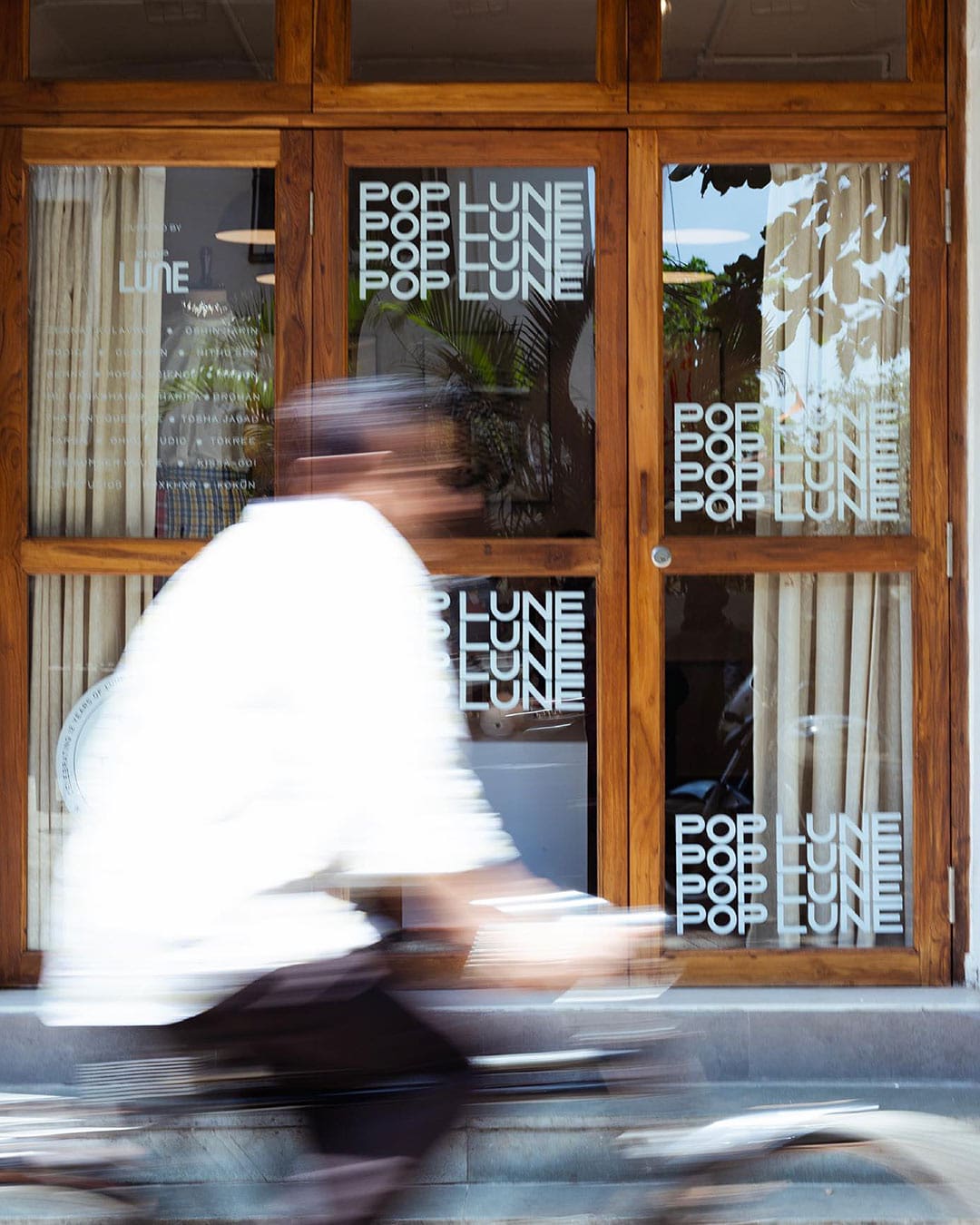 A person wearing a white shirt on a bicycle blurs past a window in a wooden frame with the words POP LUNE written in white on the glass.