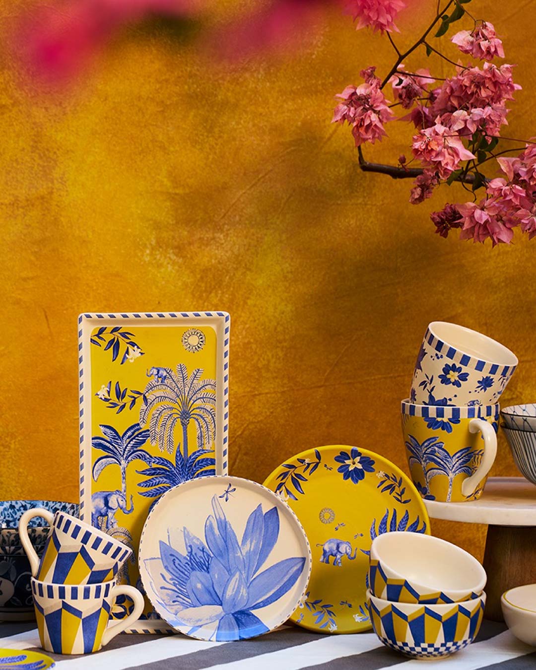 Painted ceramic mugs, plates and bowls stacked in front of an ochre wall with a pink flowering branch in the top right corner, in Nicobar boutique in Mumbai.