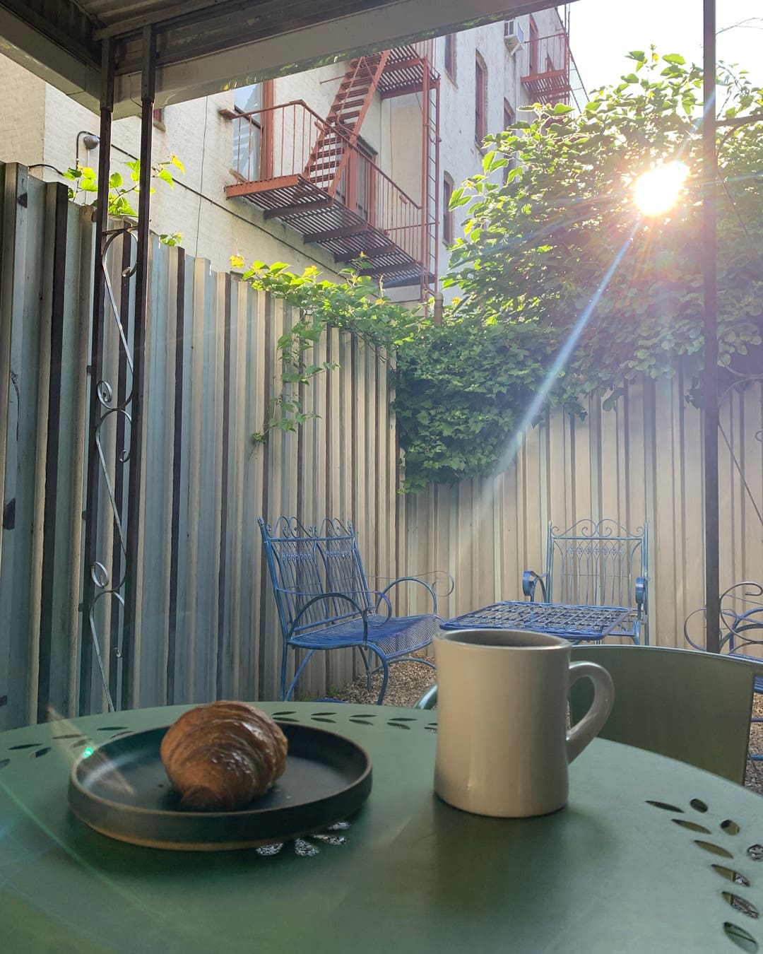 A white mug of coffee next to a croissant on a black plate on a green metal table under a shelter in the outdoor terrace area of Hamlet Coffee Company in New York.