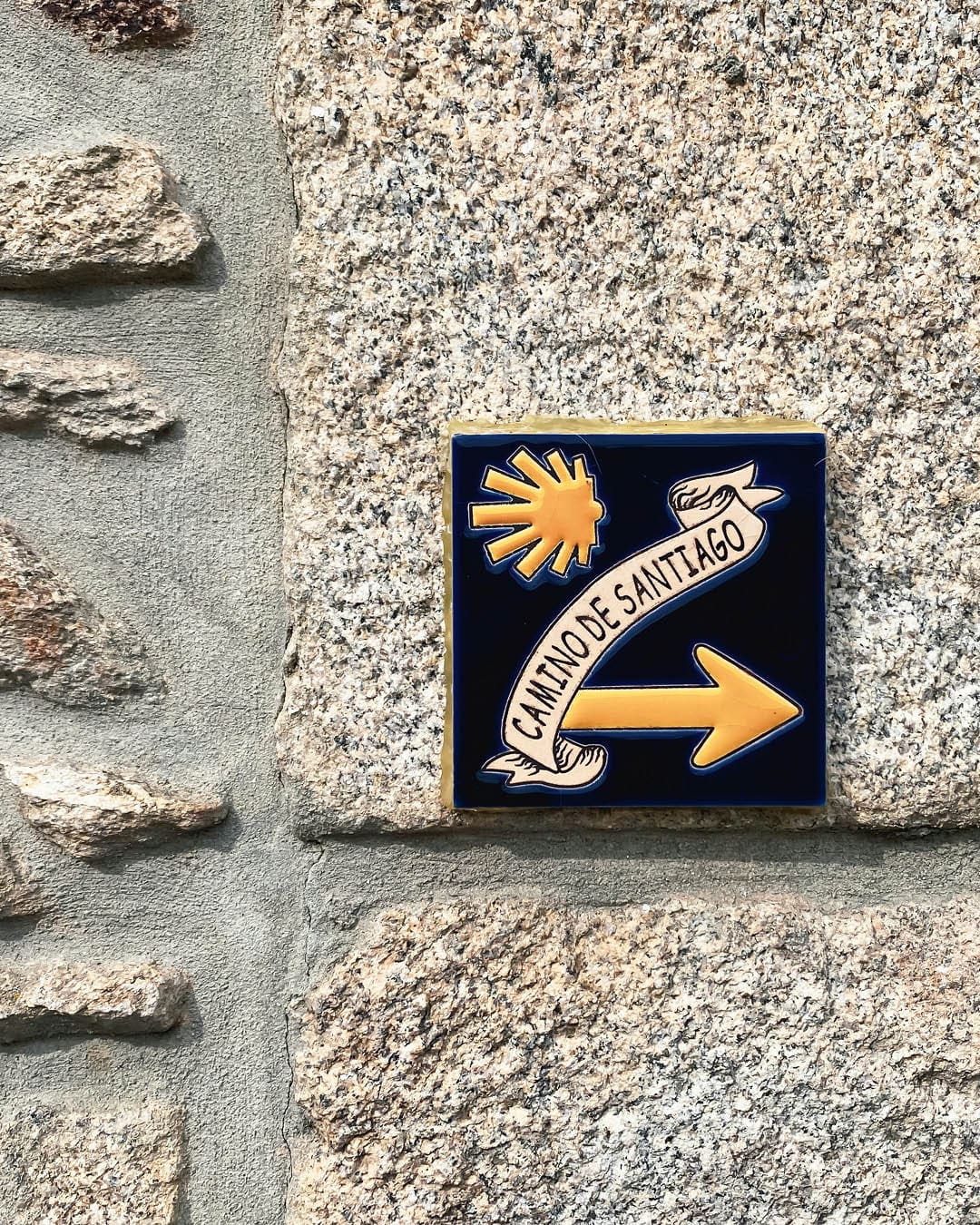 A ceramic sign with the words 'Camino de Santiago' written on it mounted to a stone wall and pointing to the right.