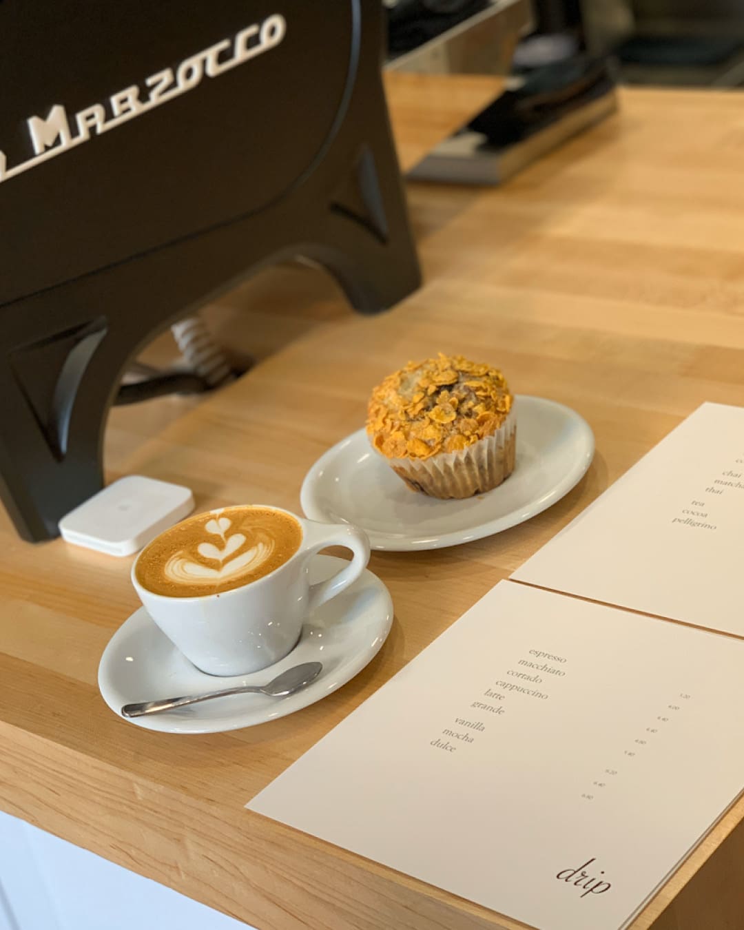 A latte and cornflake muffin served at Drip Coffee Makers on a wooden countertop next to printed menus and a black coffee machine.