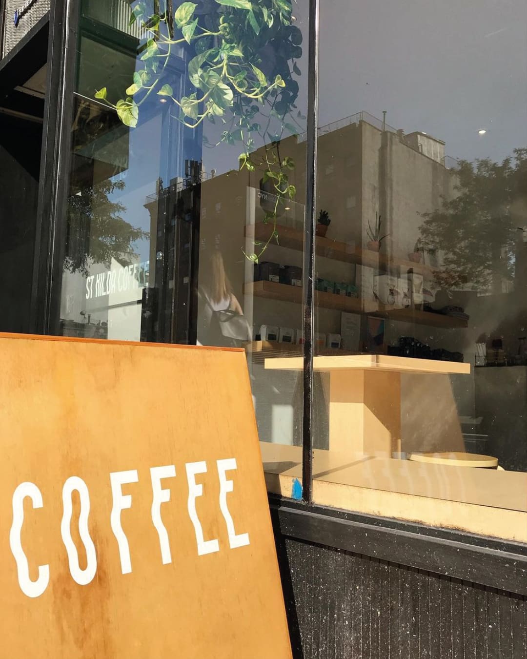 A wooden sign reading 'COFFEE' in white wobbly text in front of a tinted glass window outside St Kilda coffee shop in New York.