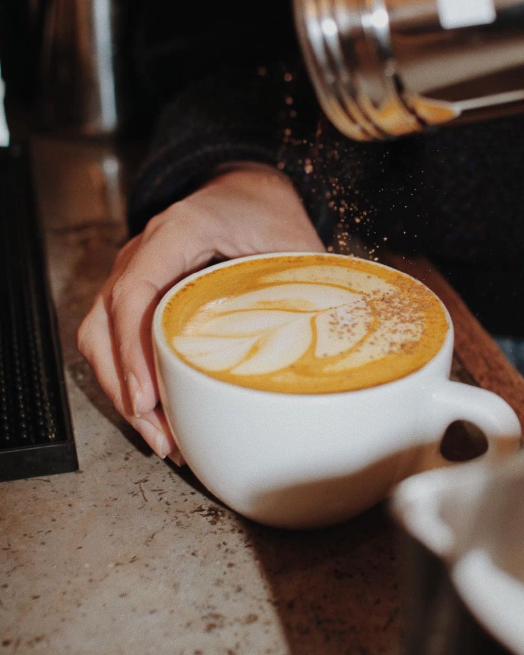 A hand holds a cappucino while a shaker dusts the top at Kinship Coffee Collective in New York.