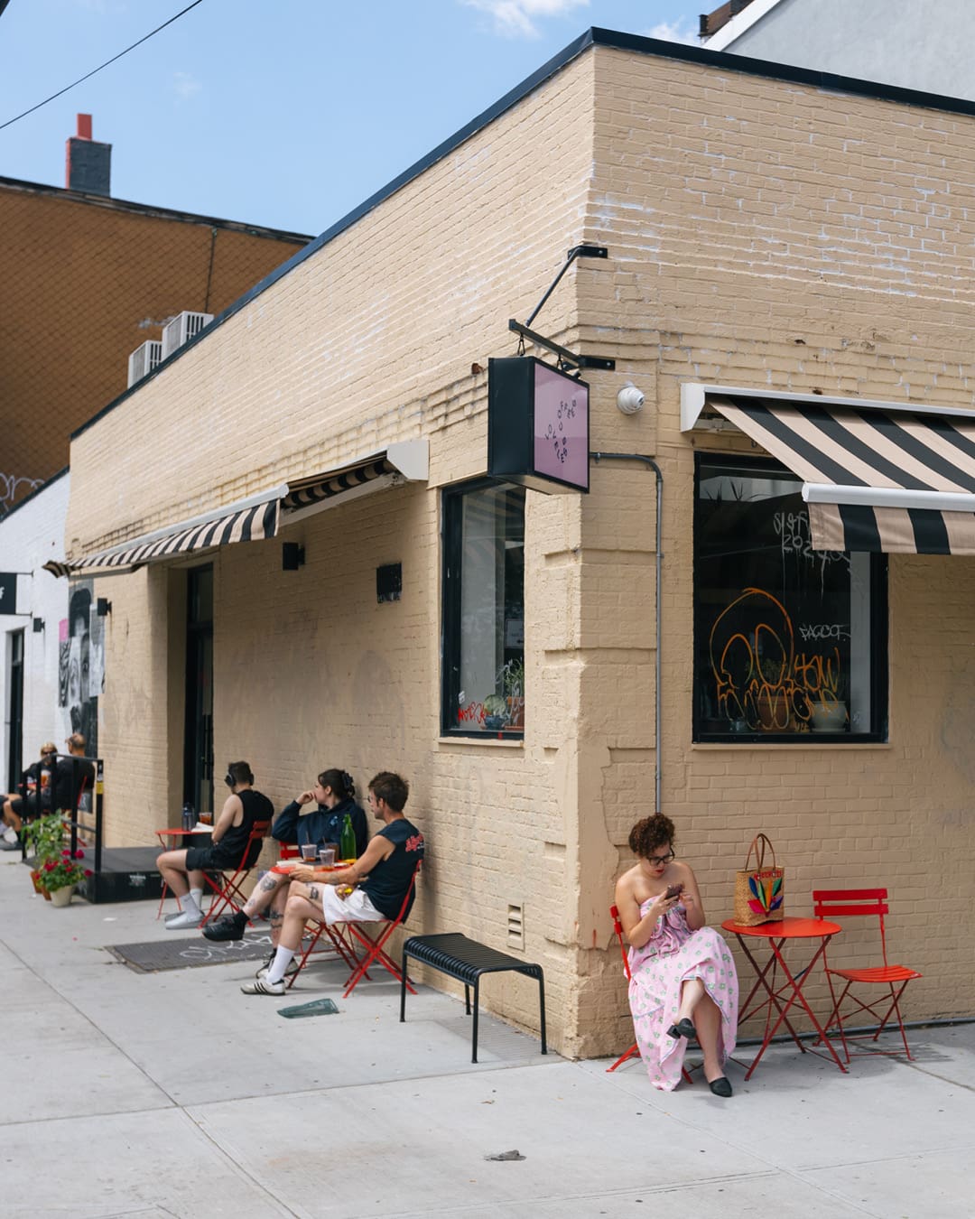 The sand-toned exterior of Loveless Coffees, on a corner in New York, with black and white striped awnings, red tables, chairs and benches and customers sat down.