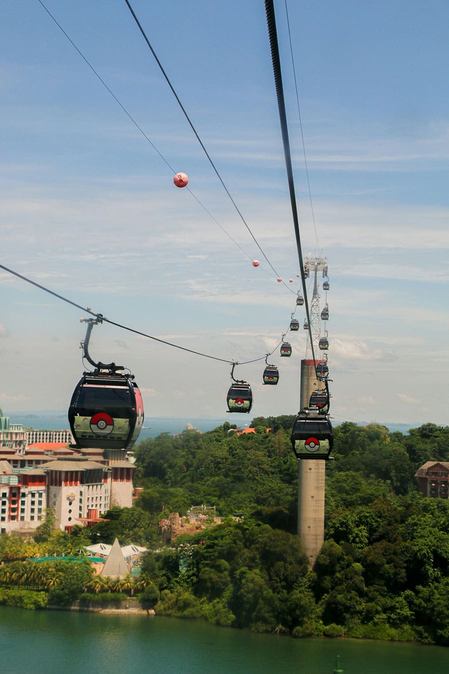 A series of cable cars travels over water, trees and buildings in Sentosa, an accessible travel destination in Singapore.