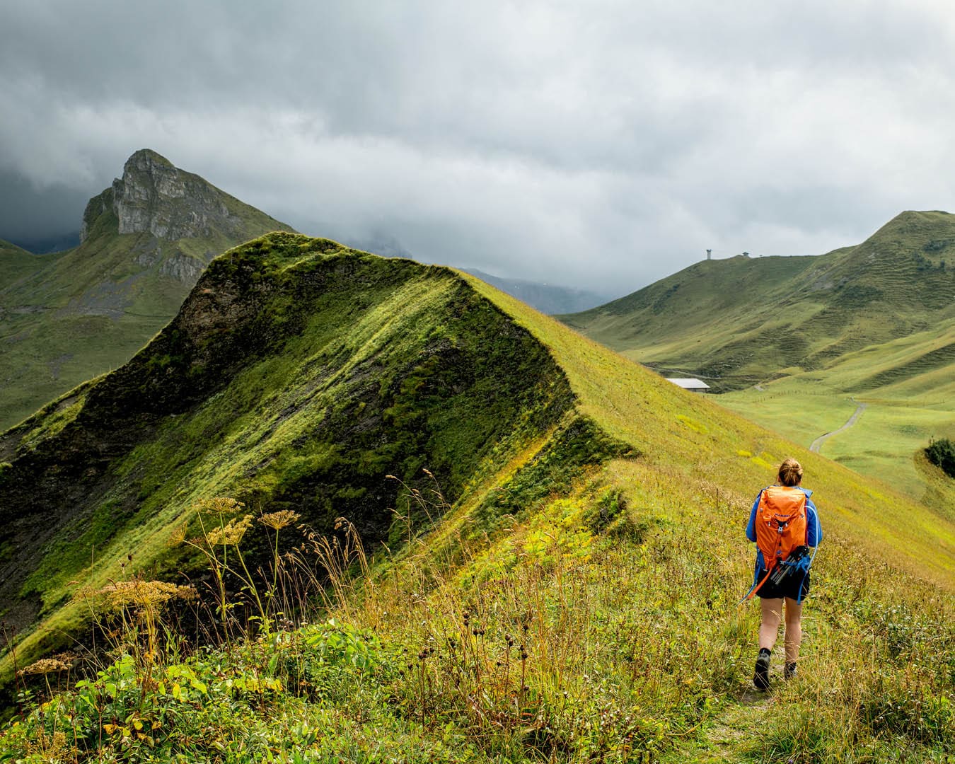 A hiker with an orange bag walks past a green mountainous ridge, with mountains in the background and a grey sky overhead.