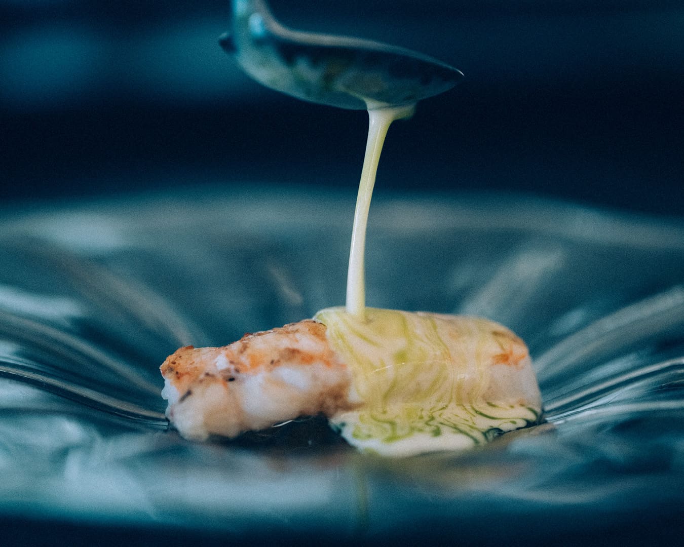 A close up of a creamy sauce being poured on a piece of seafood at the centre of a large dark blue bowl.