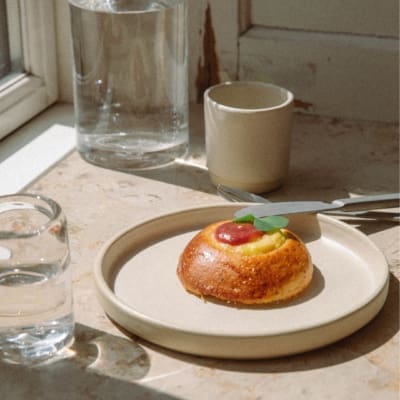 A sourdough bun on a beige plate, with a glass of water, a mug, and a jug on a marble windowsill.