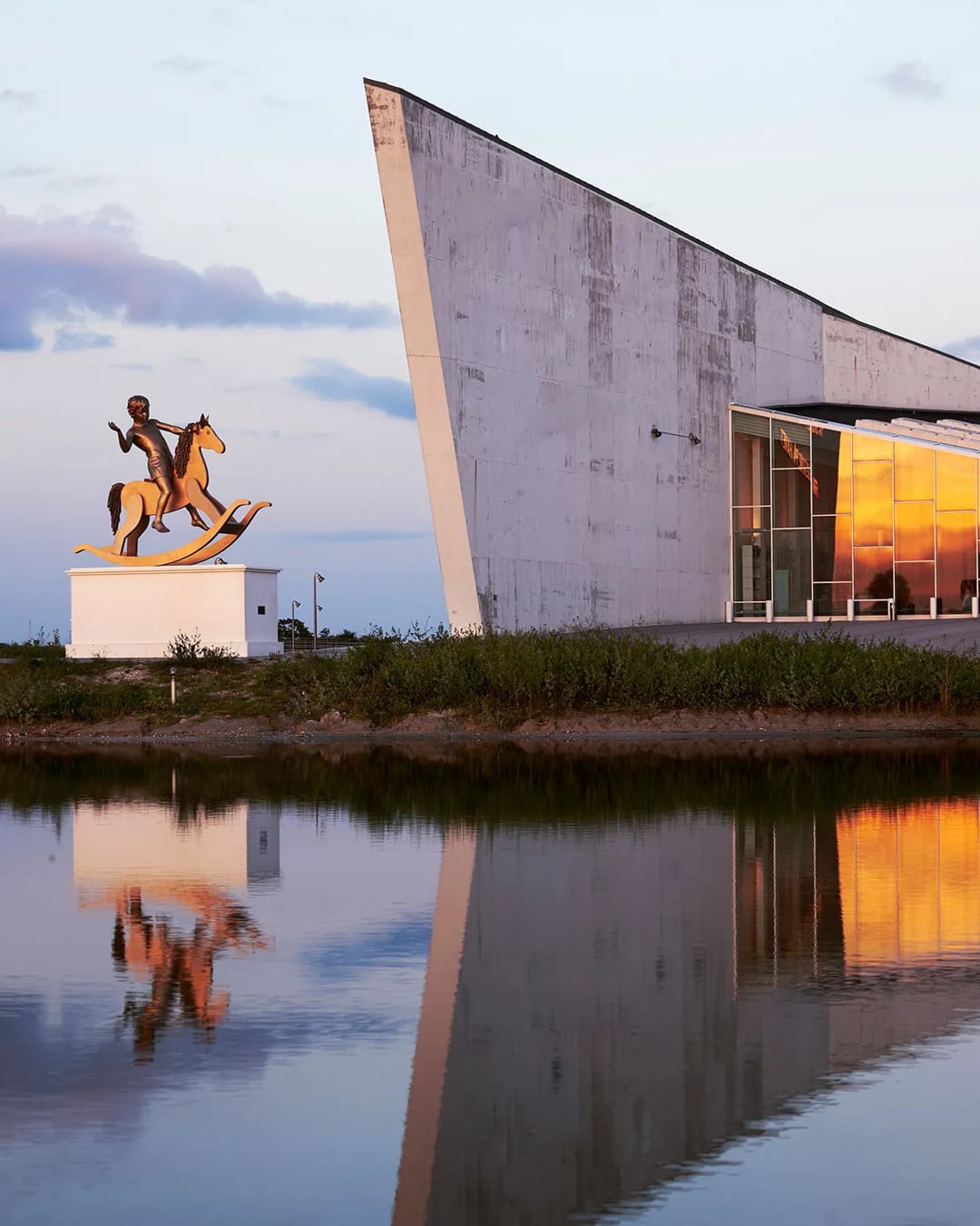 The angular facade of ARKEN Museum of Contemporary Art, reflected in water, with a sculpture outside.