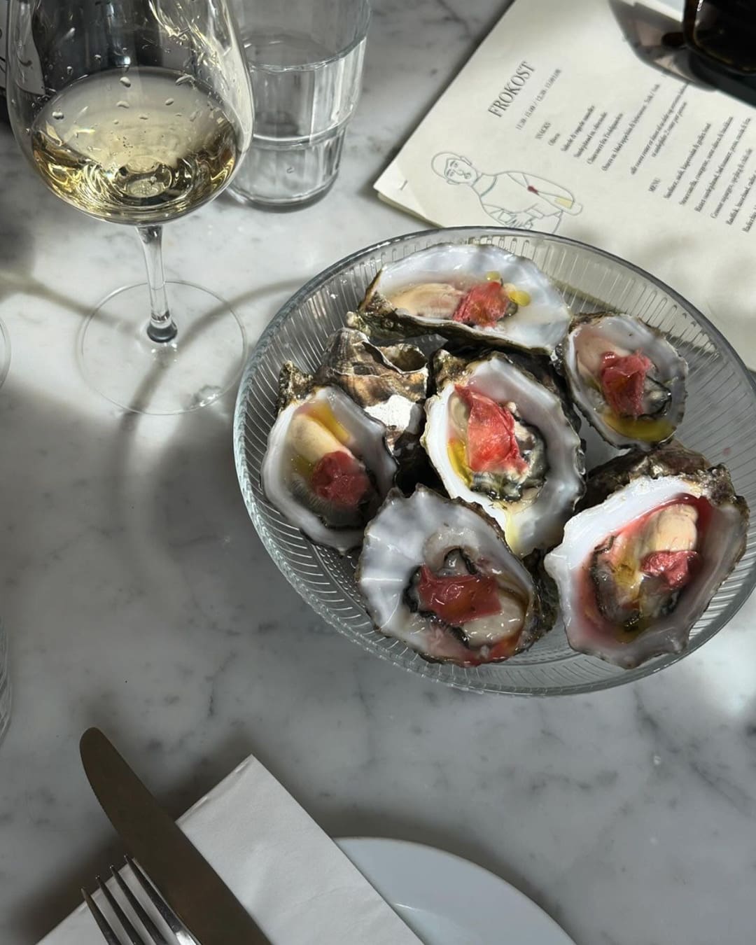 A bowl of oysters next to a glass of white wine on a marble tabletop at Bottega Barlie, Copenhagen.