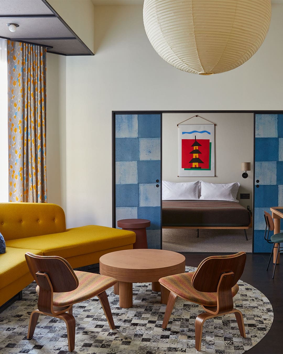 A yellow sofa sits beside two low wooden lounge chairs on a circular matt, with a bedroom seen behind through sliding doors, in Ace Hotel Kyoto.