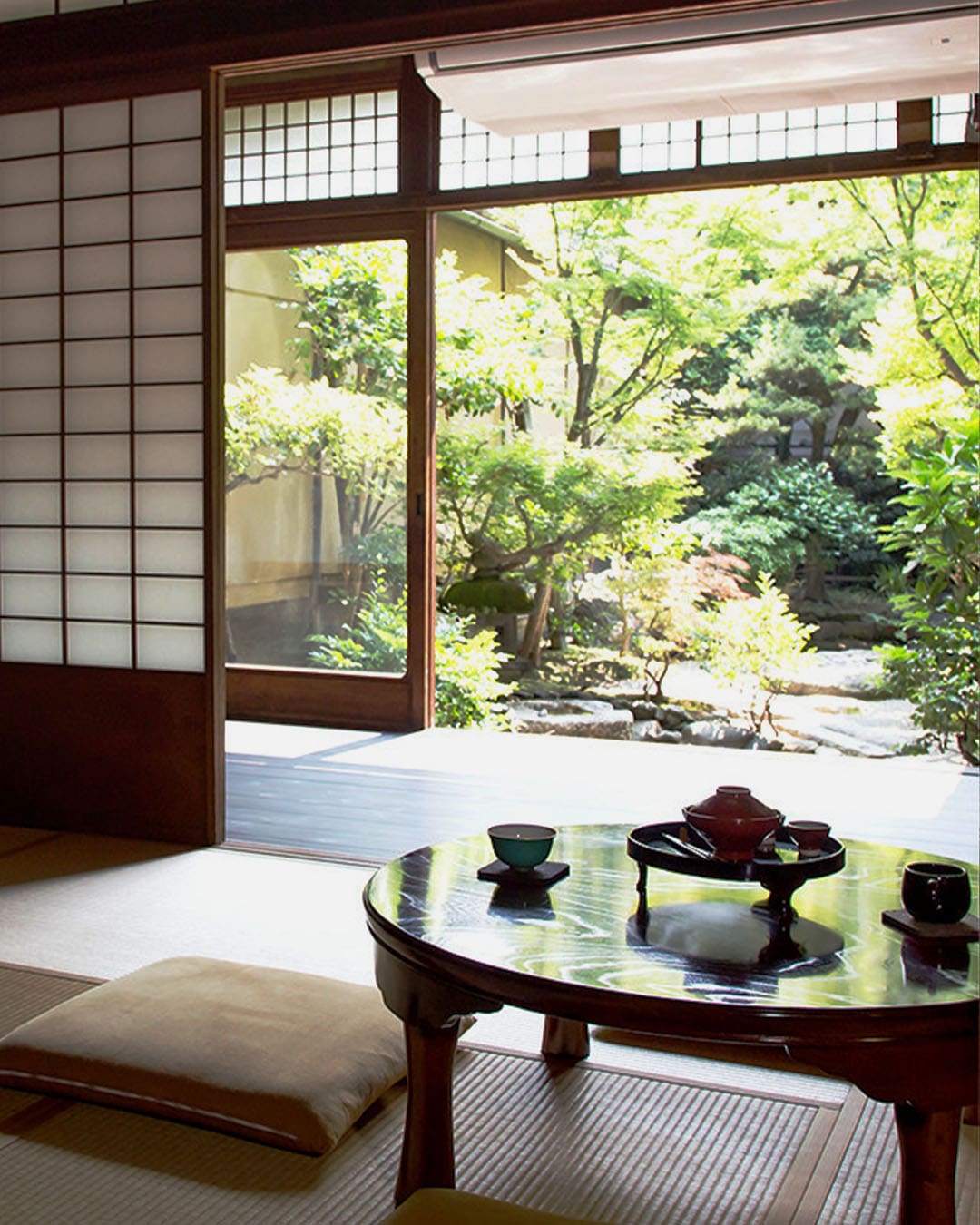 A traditional tatami room in The Terminal, housed in a machiya home. A low table with tea cups on is surrounded by cushions on a tatami reed floor, with washi screen doors open in the background to a leafy courtyard garden.