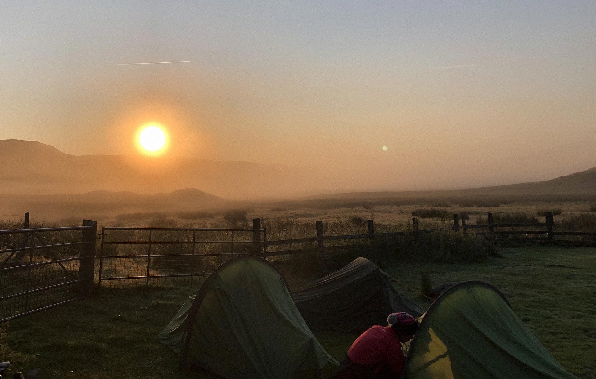 A sun rises over a misty landscape, with three small tents and a wooden fence in the foregound.