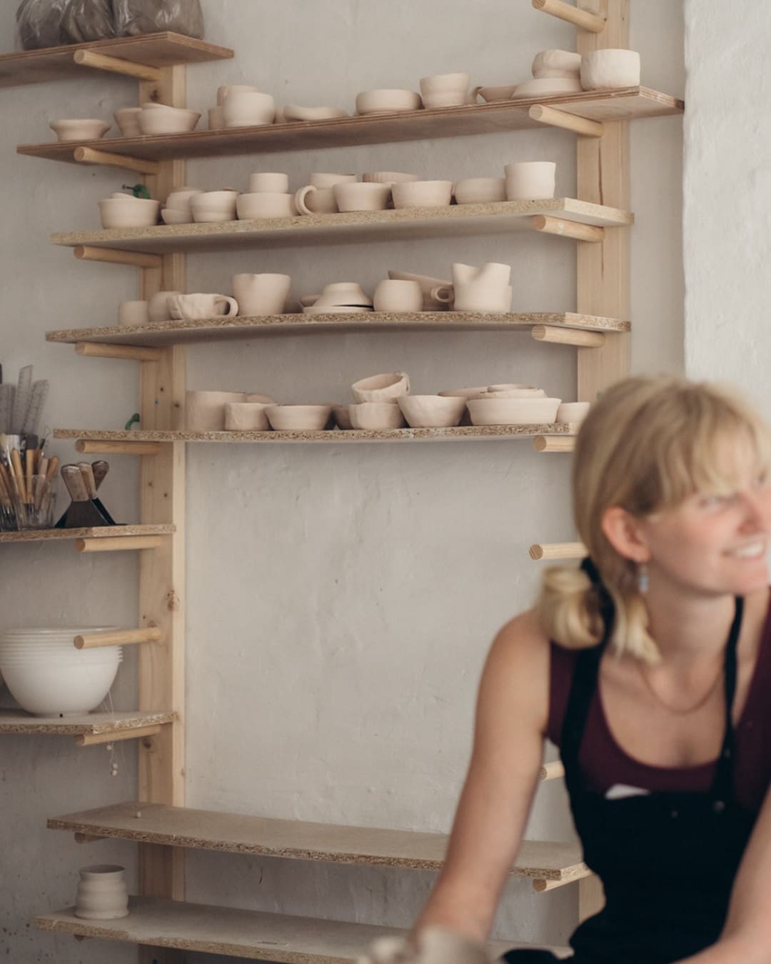 A ceramicist works in the Yōnobi workshop in Copenhagen, with drying ceramics on a shelf behind her.