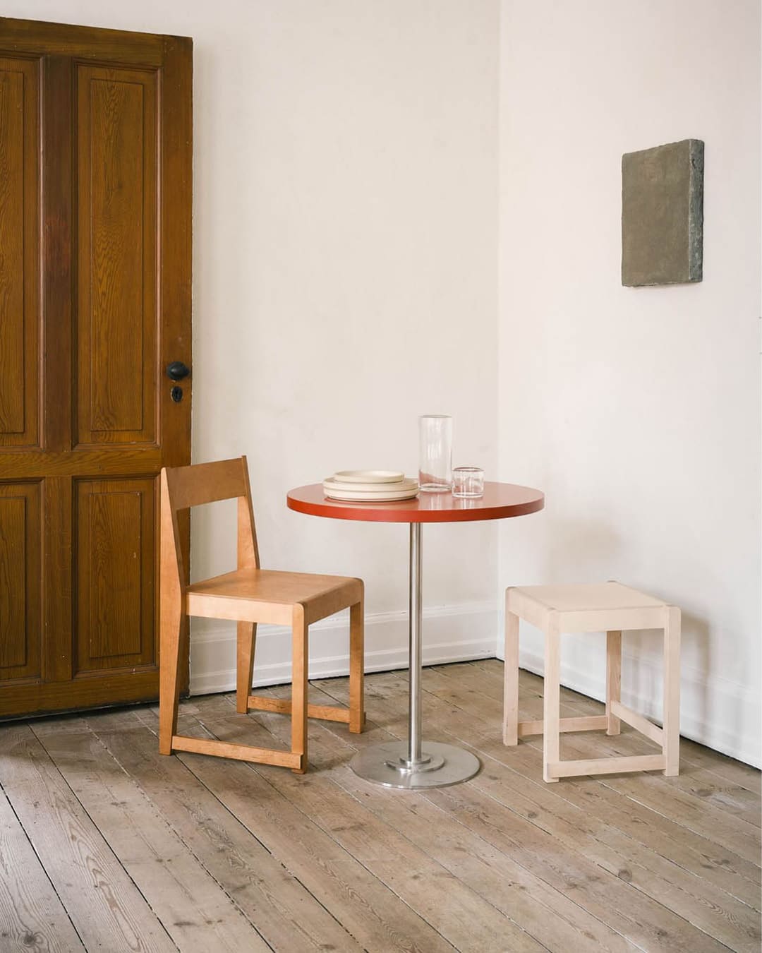 A wooden chair and stool around a red table in a white-painted room with a wooden floor, wooden door, and a small green painting on the wall at Apotek 57 café in Copenhagen.