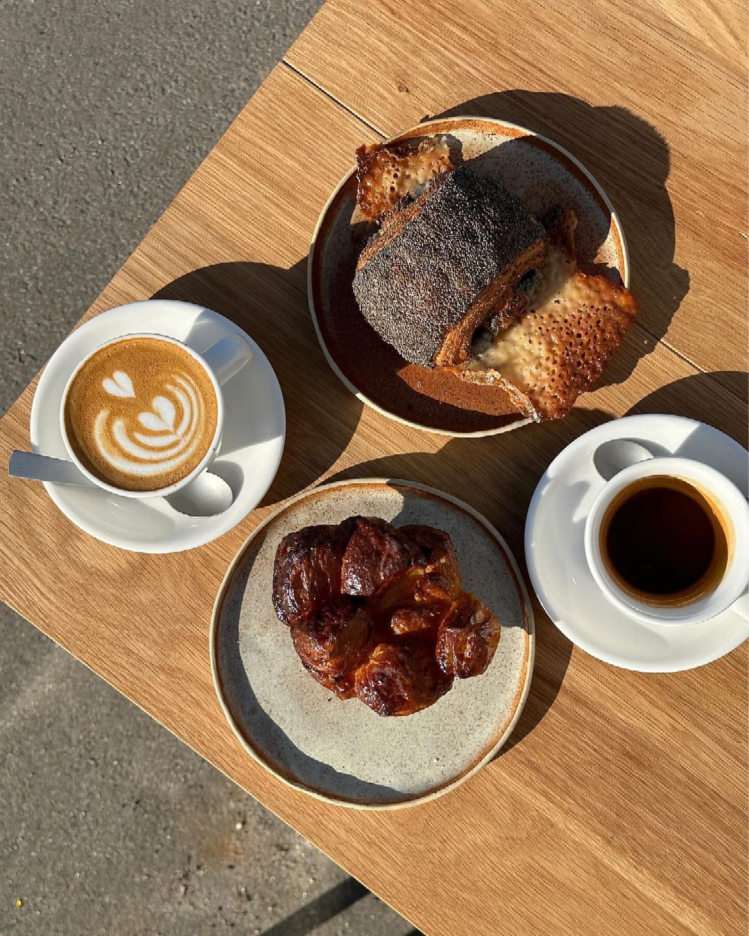 A cardamom croissant and cheese and poppy seed pain served with a flat white coffee and a black coffee on an outdoor wooden tabletop outside Hart Bageri in Copenhagen.