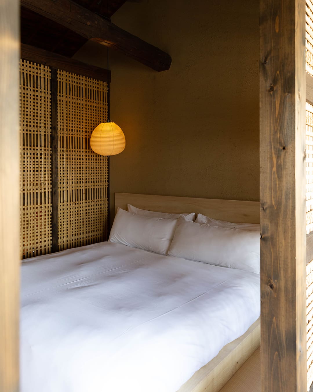 A shoji paper lamp hangs over a double bed made up with white sheets in a small wooden room with a woven screen and wood panelling at Maana Kiyomizu in Kyoto.