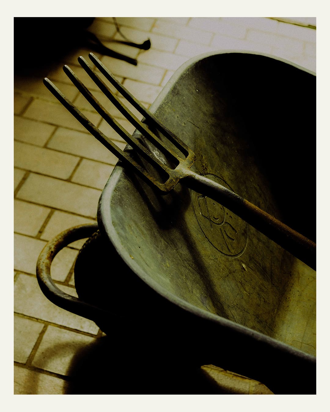 A detail of a green pitchfork resting on a green wheelbarrow above a floor of yellow tiles