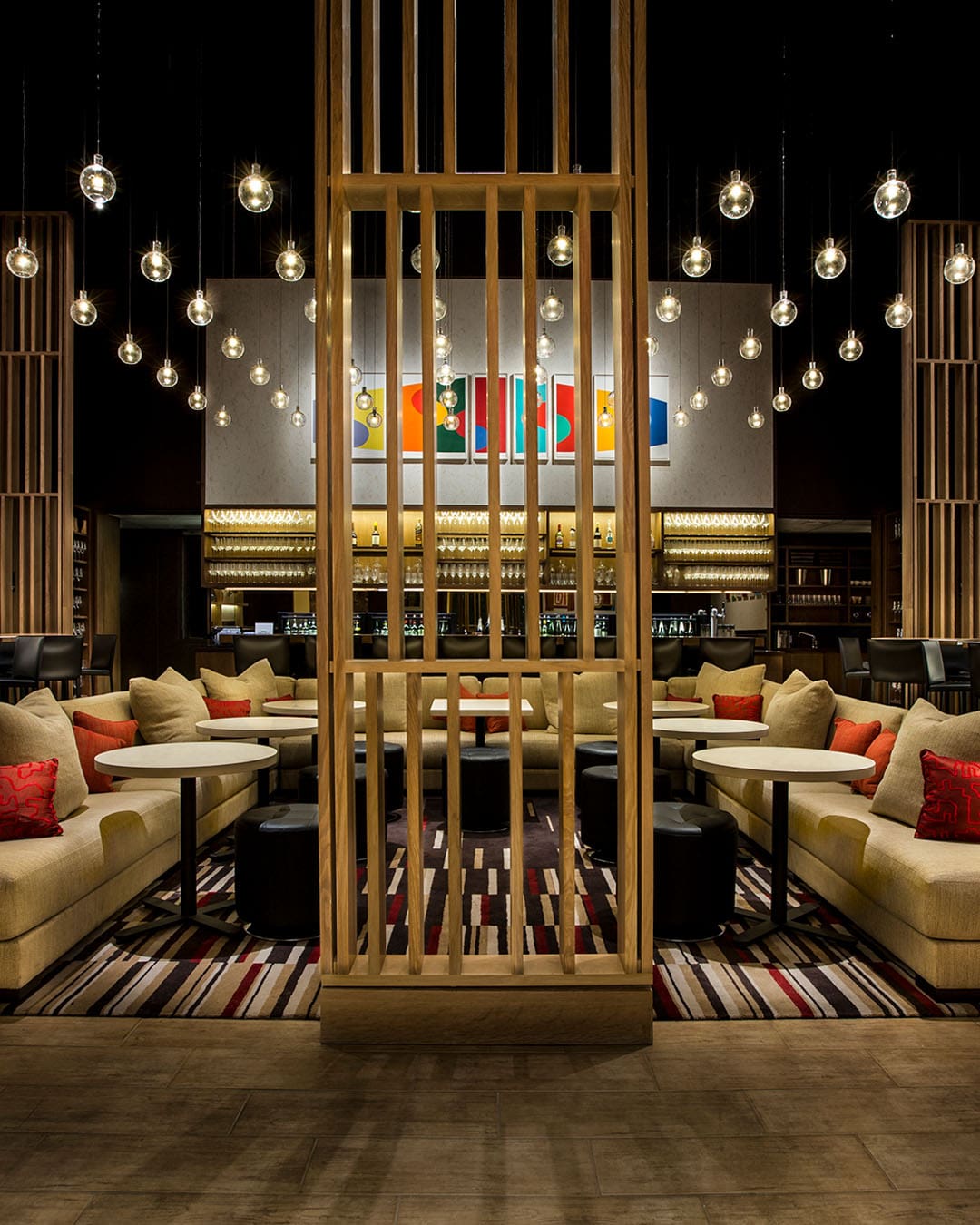 A large couch with pillows, individual tables with round ottomans and tall bar stools, under hanging lights with eclectic pieces of art in front of the fully stocked bar of the Aldo Sohm Wine Bar, NYC