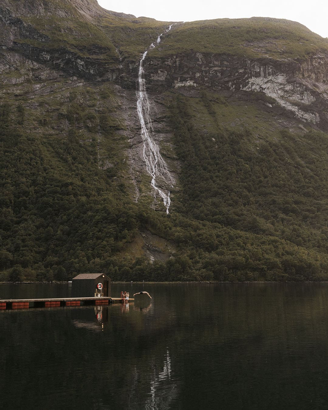 A person dives from the dock at Hotel Union Øye, Norway. Background of a lake and mountain with stream with waterfall
