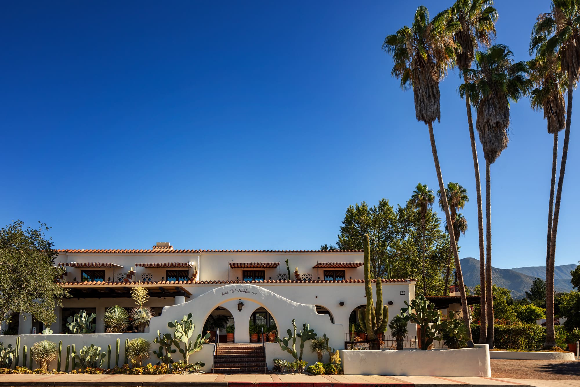 The white Mission Revival exterior of Hotel El Roblar beneath a blue sky