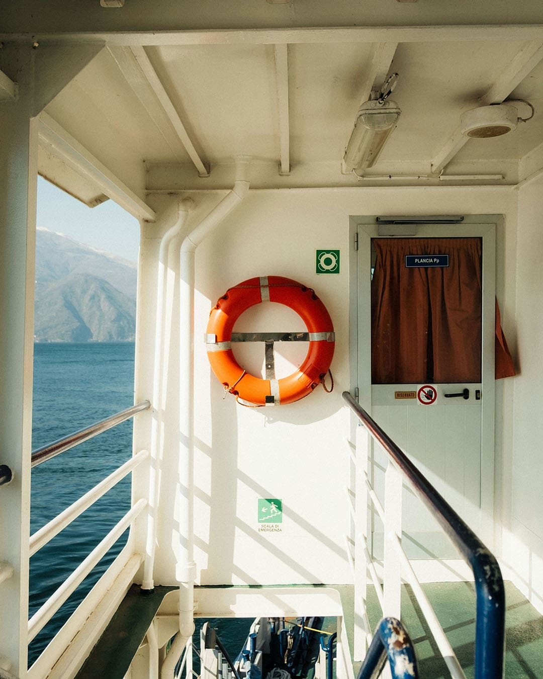 Covered deck of a ferry on Lake Como with life buoy mounted above a companionway and a glimpse of lake and mountain over side railings