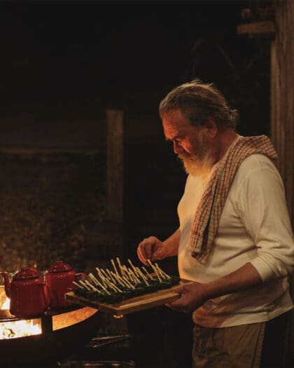 Bearded chef prepares fresh food dishes over open flames with guests in background, atmospheric scene.