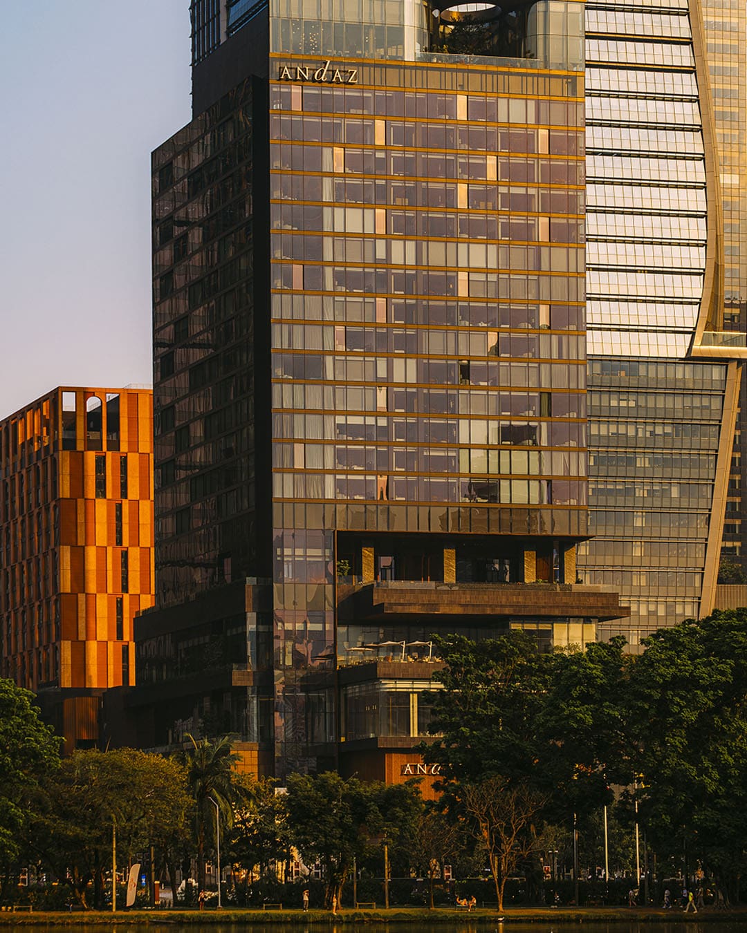 Evening sun catching the glass exterior of a modern luxury boutique hotel Andaz Bangkok. Framed by greenery