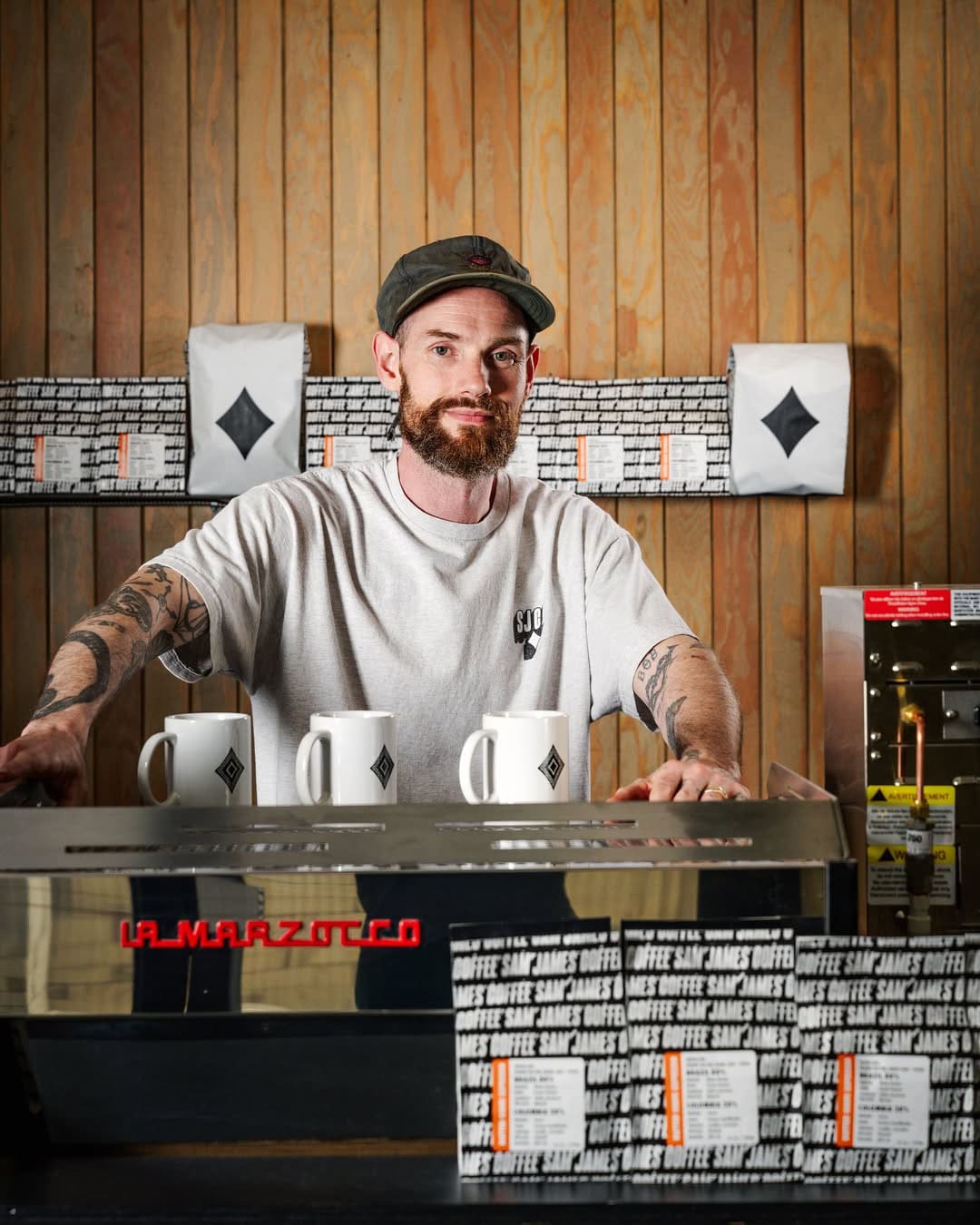 The founder of Sam James Coffee Bar poses for a photograph in his establishment, with coffee machine in foreground